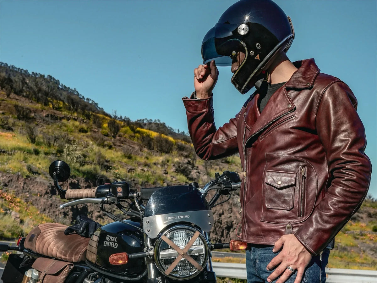 Person wearing a leather jacket and helmet next to a motorcycle with a scenic background