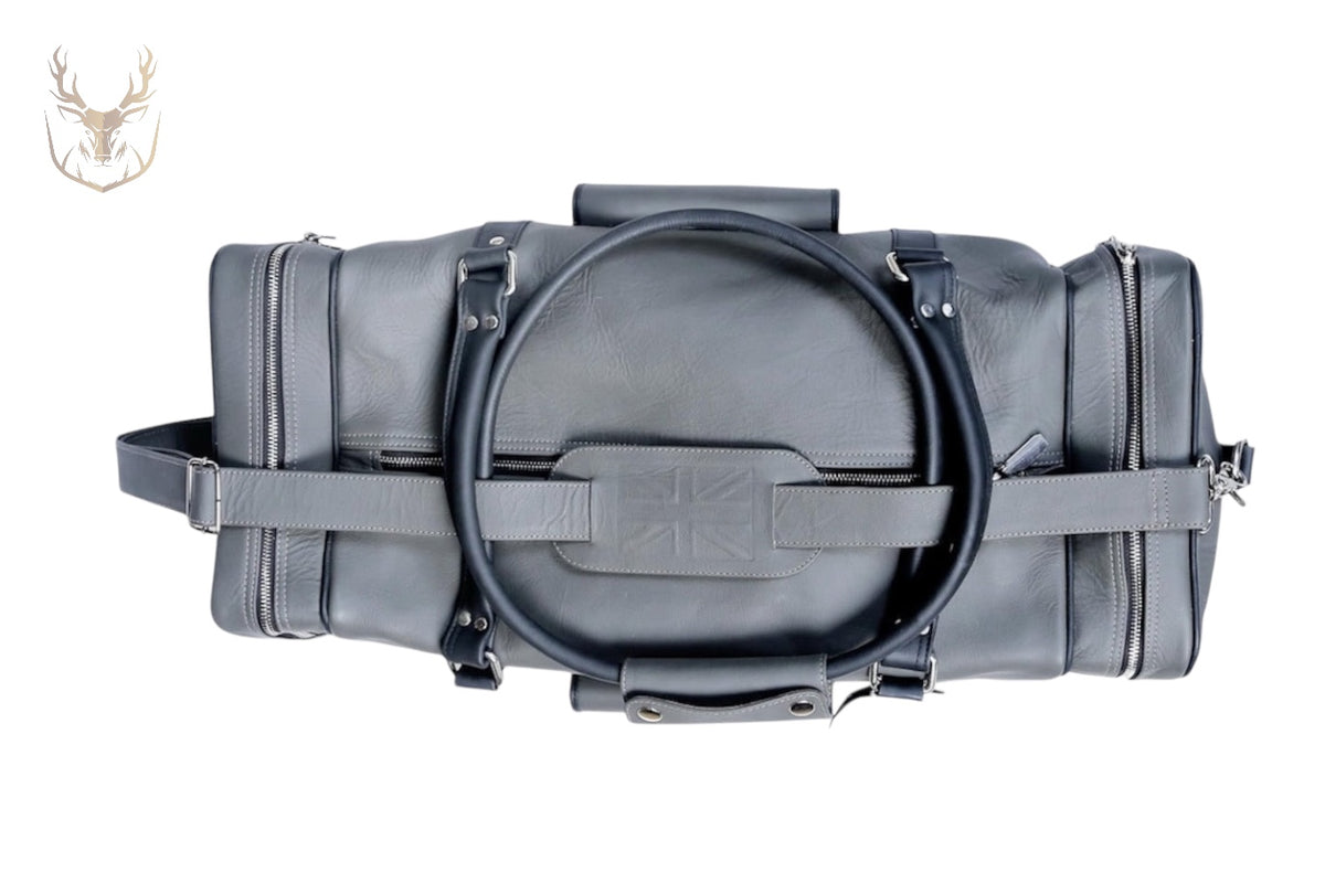 A top view of LuxeAura’s Gray Handcrafted Italian Leather Duffle Bag, featuring black handles, silver zippers, a subtle embossed Union Jack on the side, and a stag head logo in the top left corner against a white background.