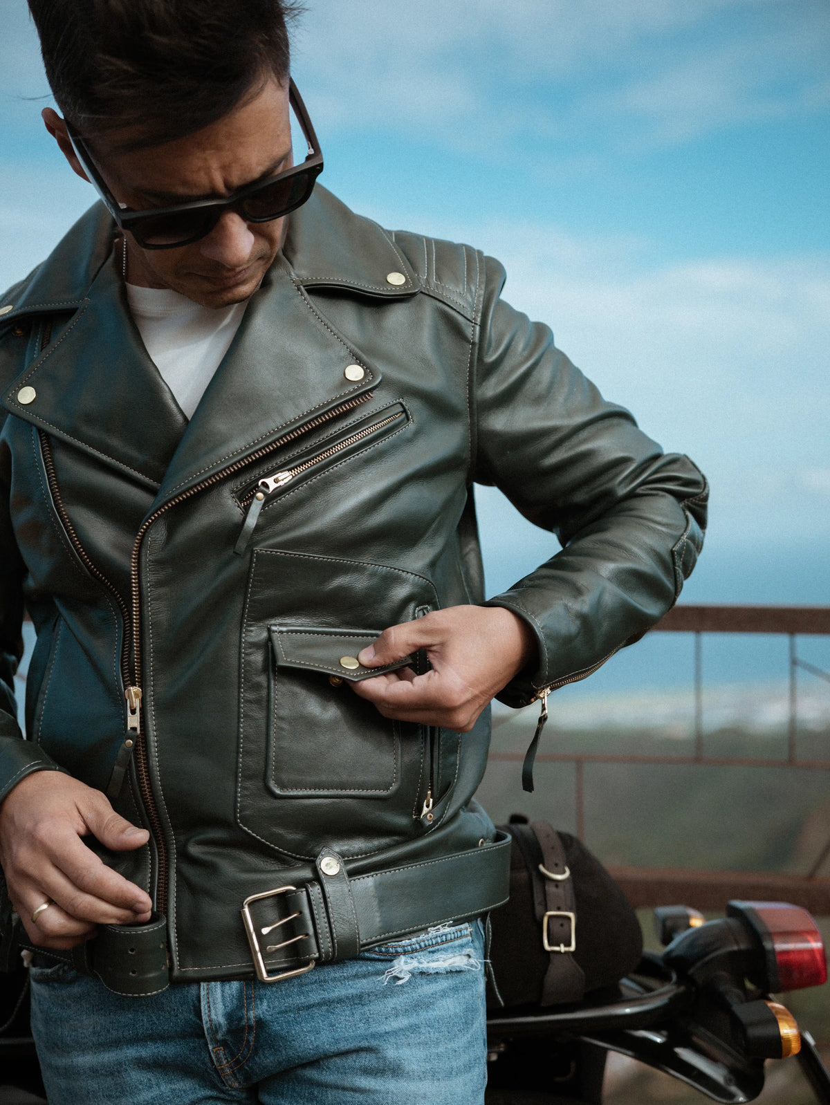 Man wearing a leather jacket and sunglasses, standing next to a motorcycle with a clear sky background.