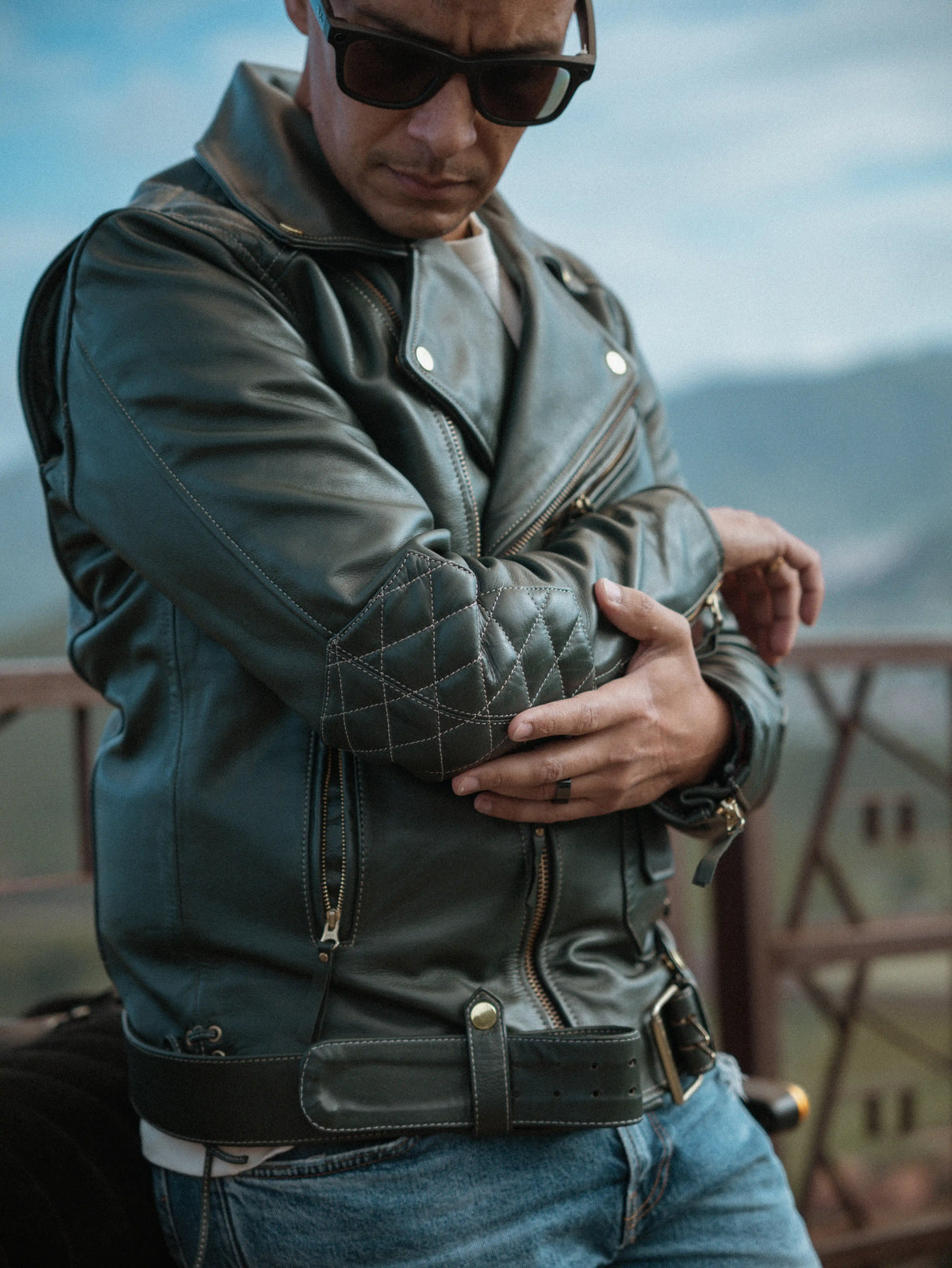 Man wearing a green leather jacket and sunglasses with a mountainous background