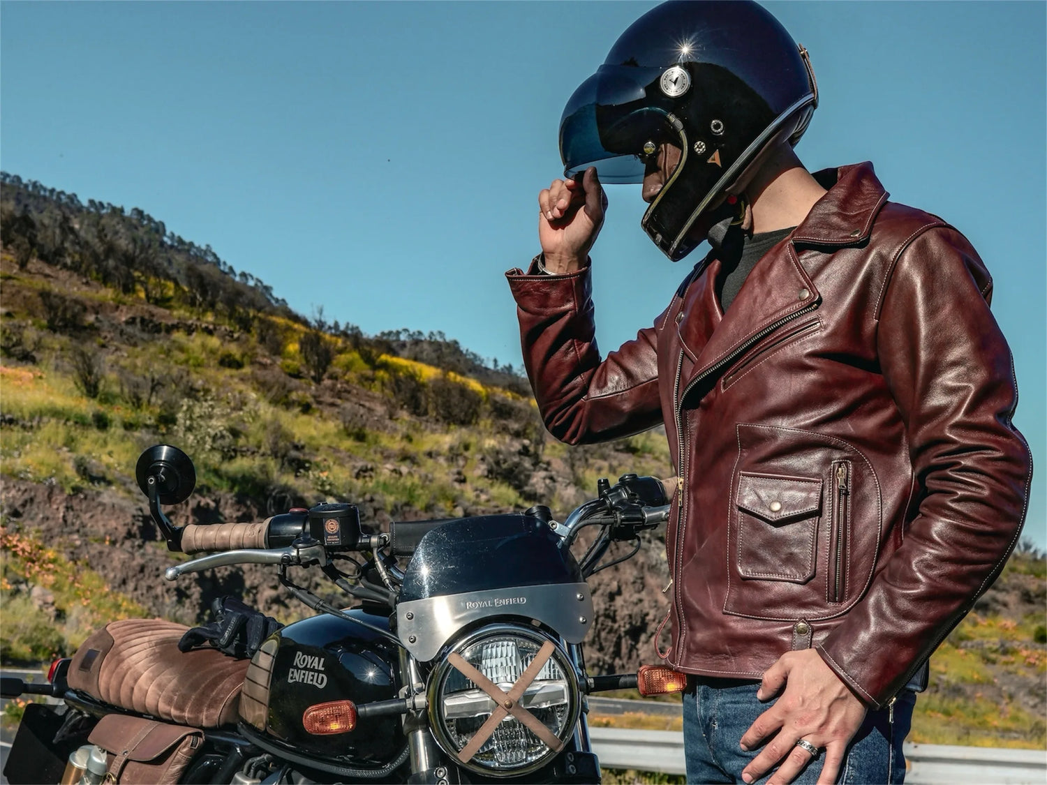 Person wearing a leather jacket and helmet next to a motorcycle on a scenic road.