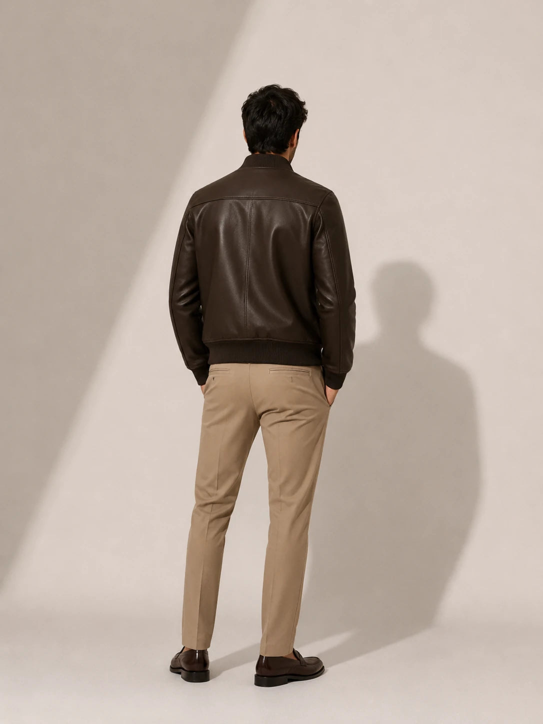 Man wearing a brown leather jacket and beige pants standing against a plain background