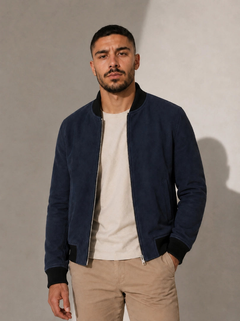 Man wearing a navy bomber jacket over a beige shirt and pants against a plain background