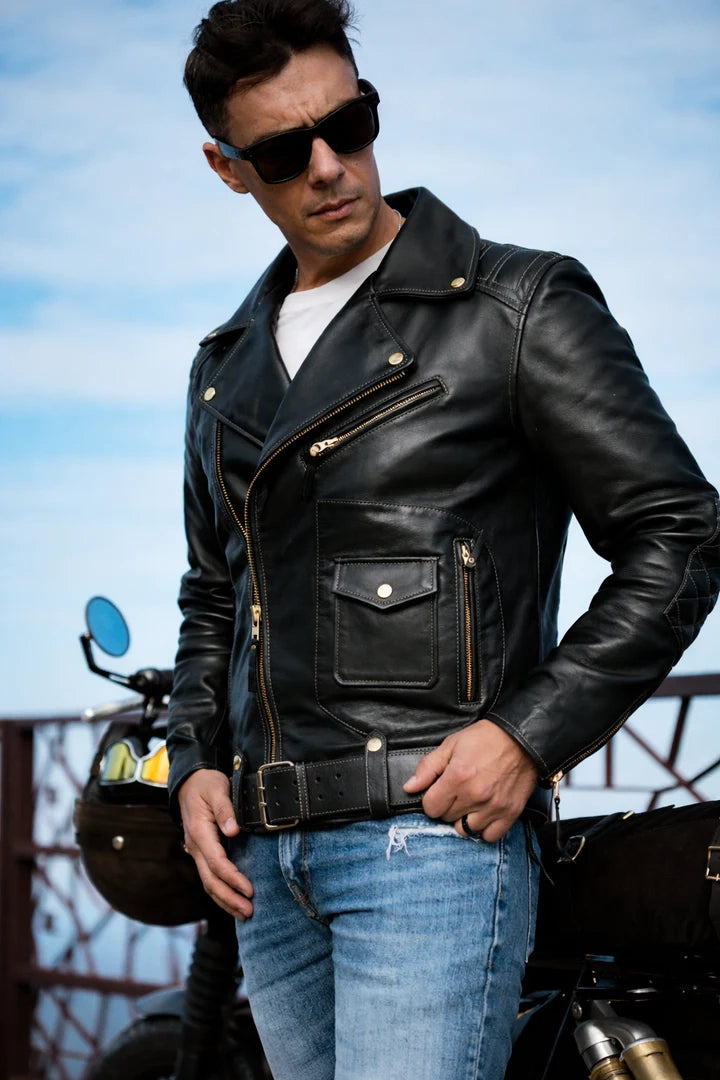 Man wearing a black leather jacket and sunglasses standing next to a motorcycle with a blue sky background.