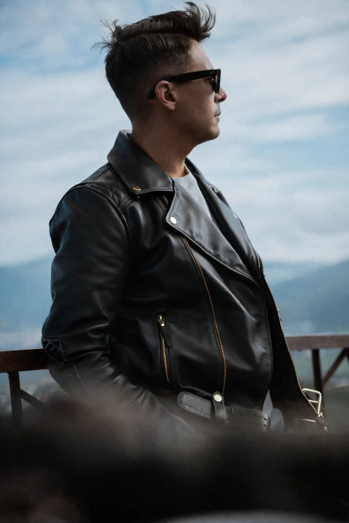 Man wearing a black leather jacket and sunglasses against a mountainous background