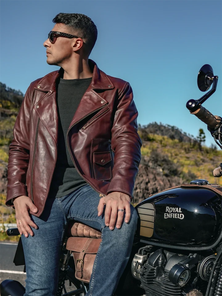 Man wearing a brown leather jacket sitting on a Royal Enfield motorcycle with a scenic background.