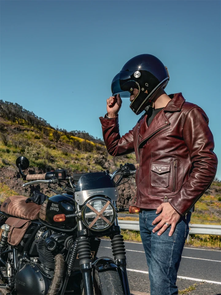 Man in a leather jacket and helmet standing next to a motorcycle on a road with a scenic background.