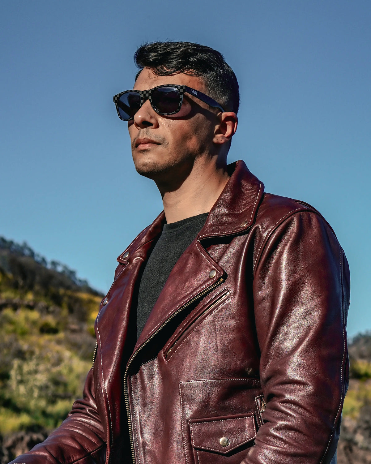 Man wearing a brown leather jacket and sunglasses with a mountainous landscape in the background